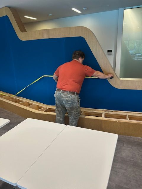 Person in orange shirt measuring a blue wall with a yellow tape measure, light wood trim, interior.
