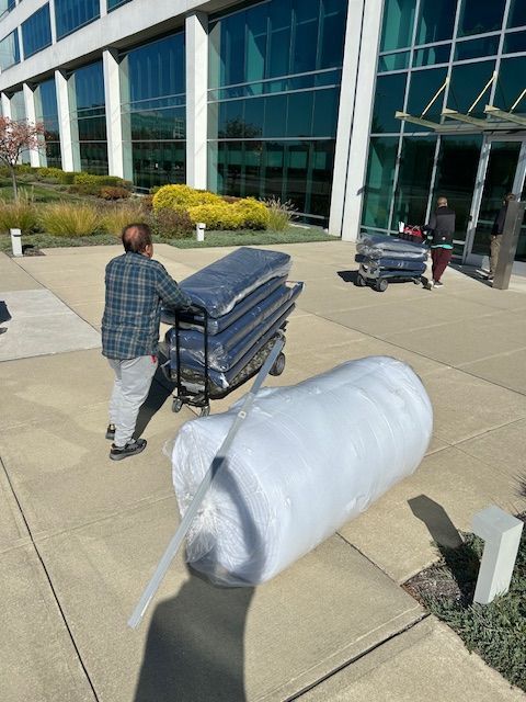 Man pushing a hand truck with stacked mattresses near a building entrance, another person in the background. A rolled mattress is on the ground.