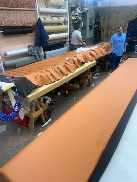 Upholstery workshop: long orange fabric piece on a work table, two men working. Rolls of fabric on the wall.