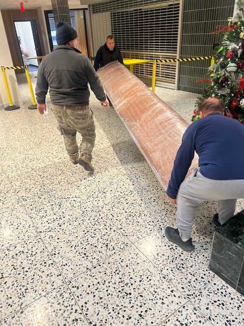 Three people carry a long, wrapped wooden object through a building lobby with a Christmas tree.