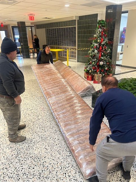 Three people move large wrapped rectangular objects in a building hallway, near a decorated Christmas tree.