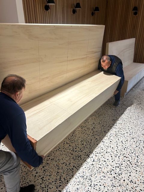 Two men carrying a large, light-colored wooden bench indoors, with a terrazzo floor.