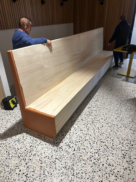 Two people installing a large, light wood bench with orange trim in a room with a terrazzo floor.