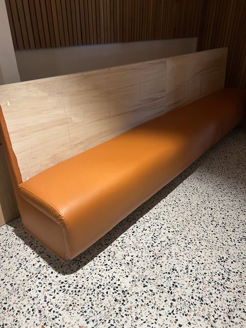 Orange leather bench seat against a light wooden backdrop, on a speckled floor.