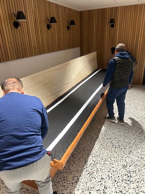 Two men carrying a long, rectangular table with a black surface and white stripes inside a room with wood paneling.