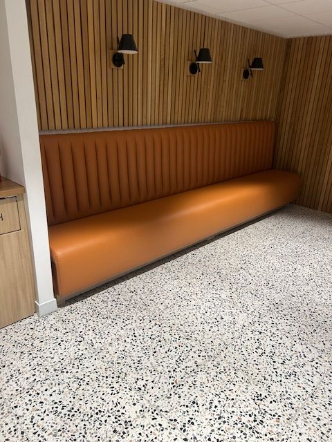 An orange booth with a ribbed back sits in a corner with wood paneling and black sconces. Speckled floor.
