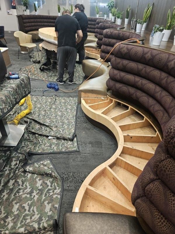 Workers reupholstering a curved booth in a commercial space. Wood frame exposed; camo carpet.