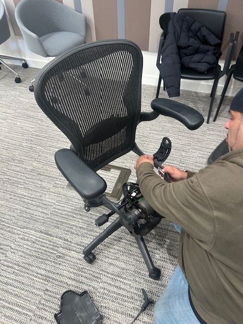 Man repairing a black mesh office chair. He's kneeling, using tools, and the chair is disassembled.