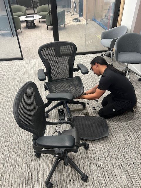 Man assembling an office chair on a patterned carpet. Office setting with other chairs visible.