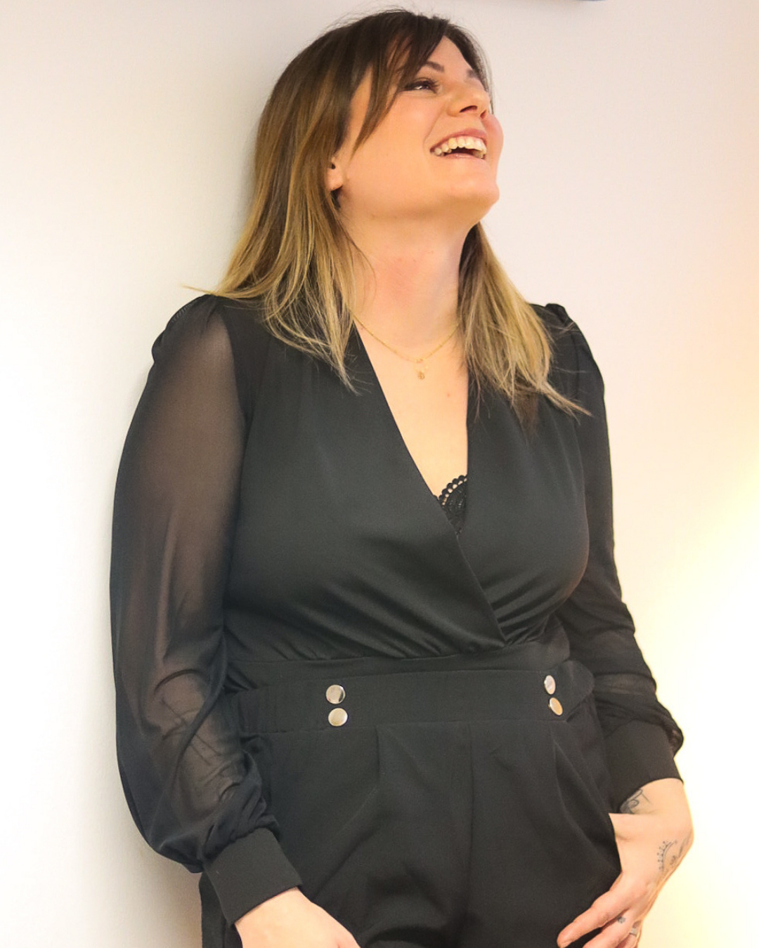 Une femme en combinaison noire rit. Elle est appuyée contre un mur blanc.