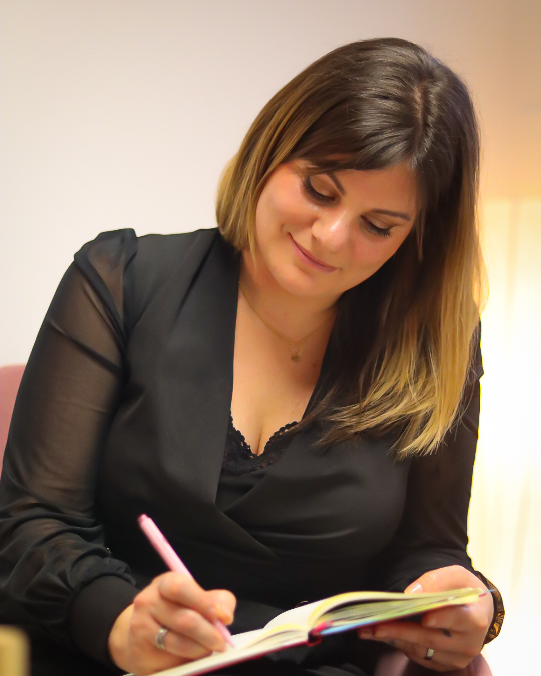 Femme en chemisier noir écrivant dans un carnet, souriant légèrement, à l'intérieur.