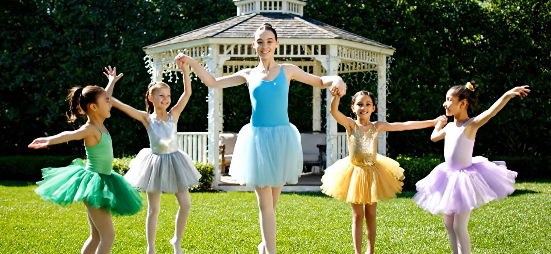 A ballet teacher with young girls, all in tutus, dancing outside. A gazebo is in the background.