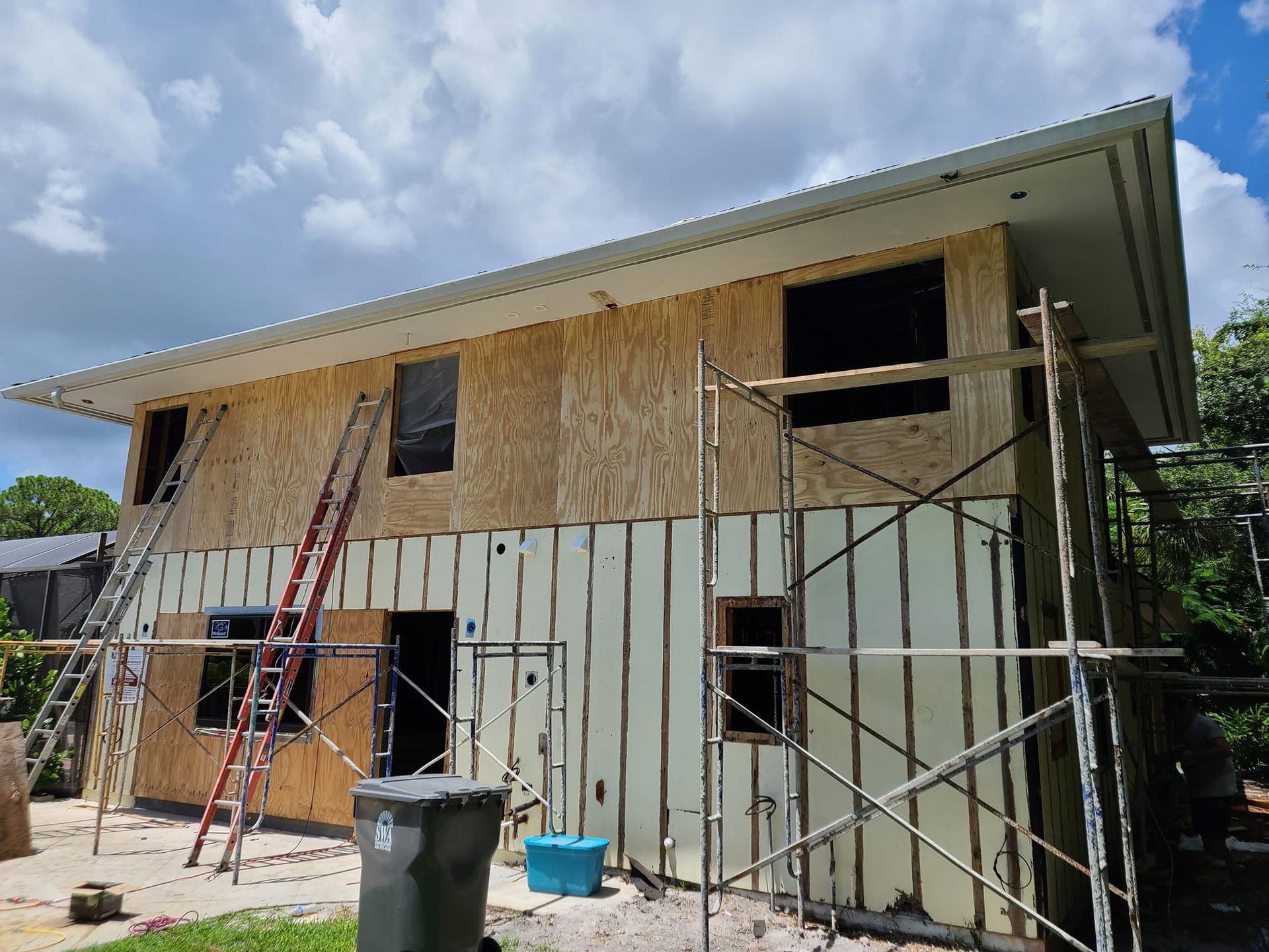 A house is being built with scaffolding and a trash can in front of it.