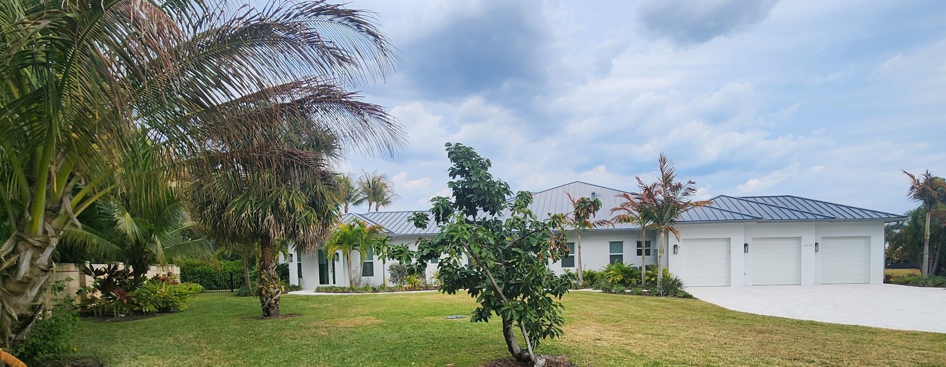 White house with three-car garage, green lawn, palm trees under cloudy sky.