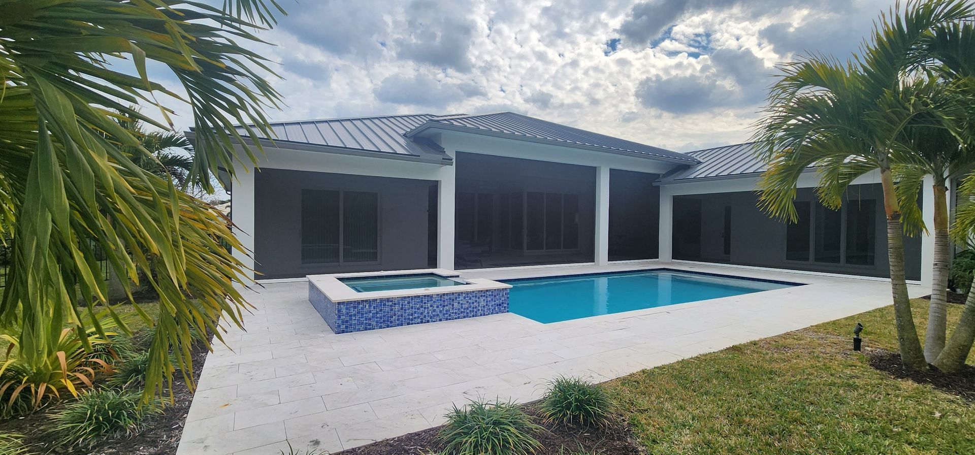 Exterior view of a house with a pool and palm trees. White patio, blue water, and gray walls.