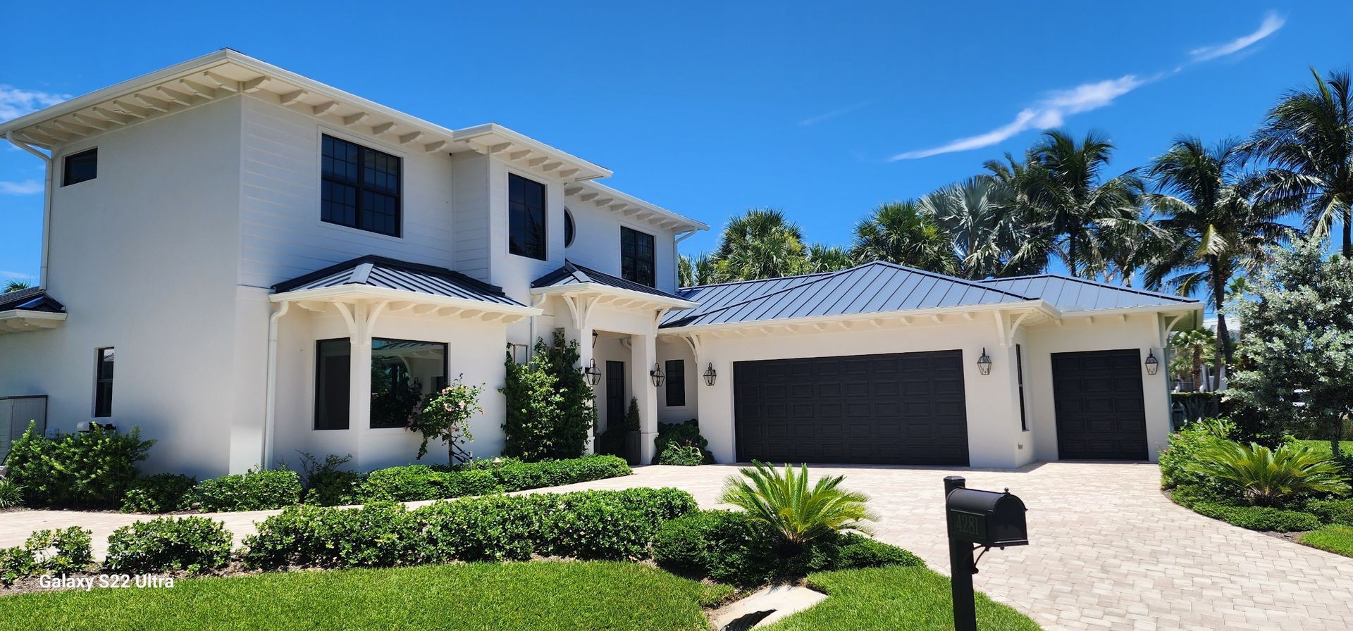 White stucco house with dark roof and garage doors, surrounded by green landscaping under a blue sky.