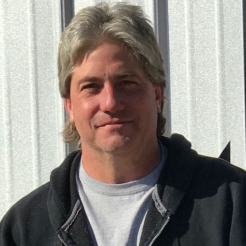 A man wearing a black jacket and a white shirt is standing in front of a white wall.