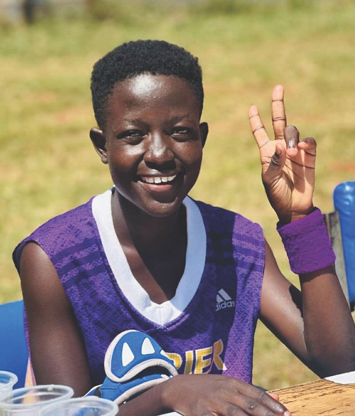 A smiling person in a purple athletic jersey makes a peace sign with their fingers while sitting outdoors.