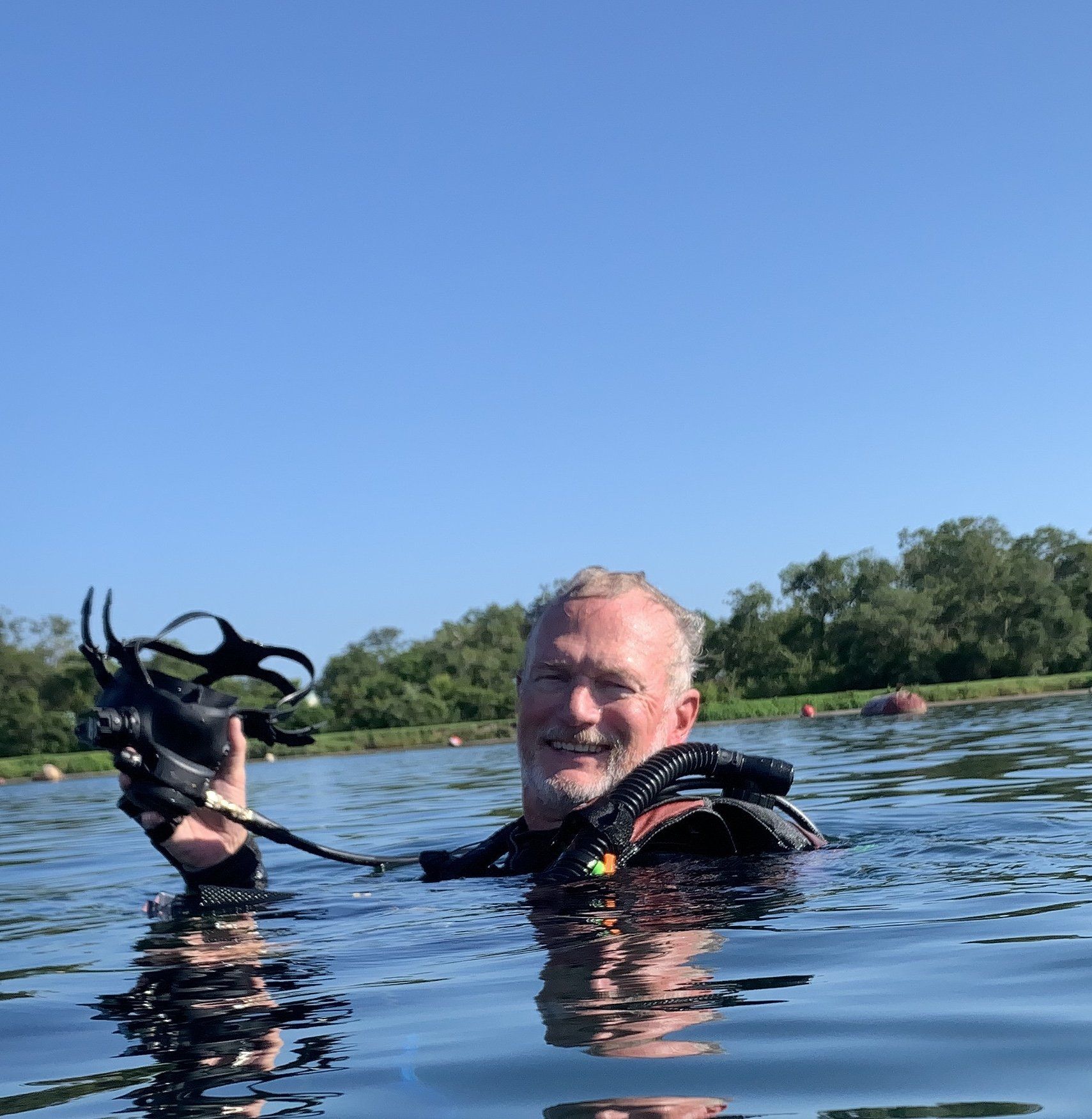 Man in scuba gear in water, holding equipment, smiling. Blue sky and trees in background.