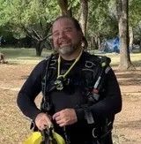 Man in diving gear smiles outdoors, trees in background.