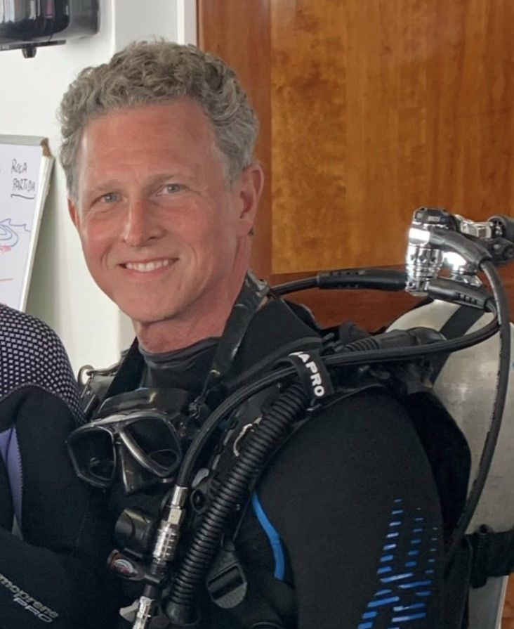 Man in diving gear smiles, indoors, next to a whiteboard.