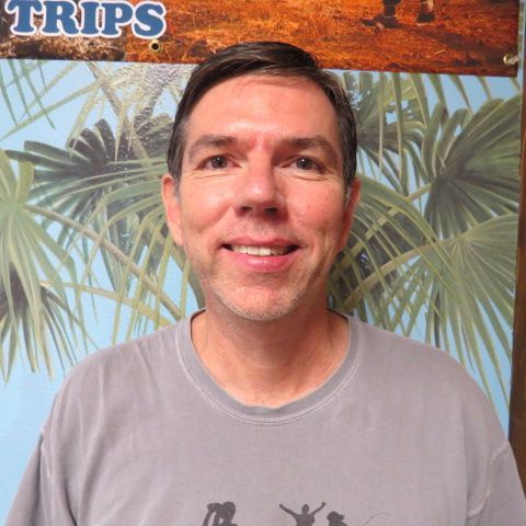 Man with short dark hair smiles, standing in front of palm tree mural.