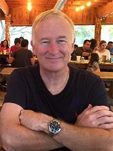Man with crossed arms, wearing watch, smiles in outdoor restaurant with people dining in background.