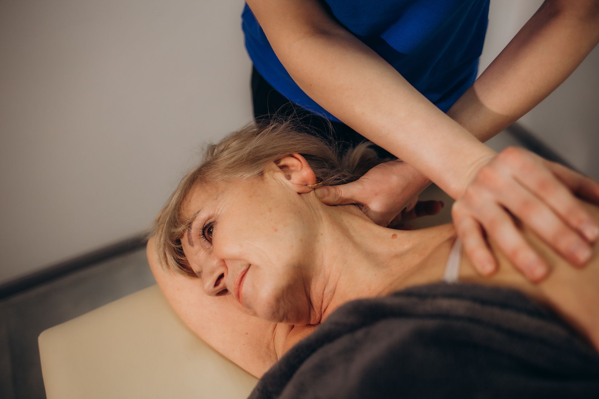 A person receiving a neck massage on a massage table. The person giving the massage wears a blue shirt.
