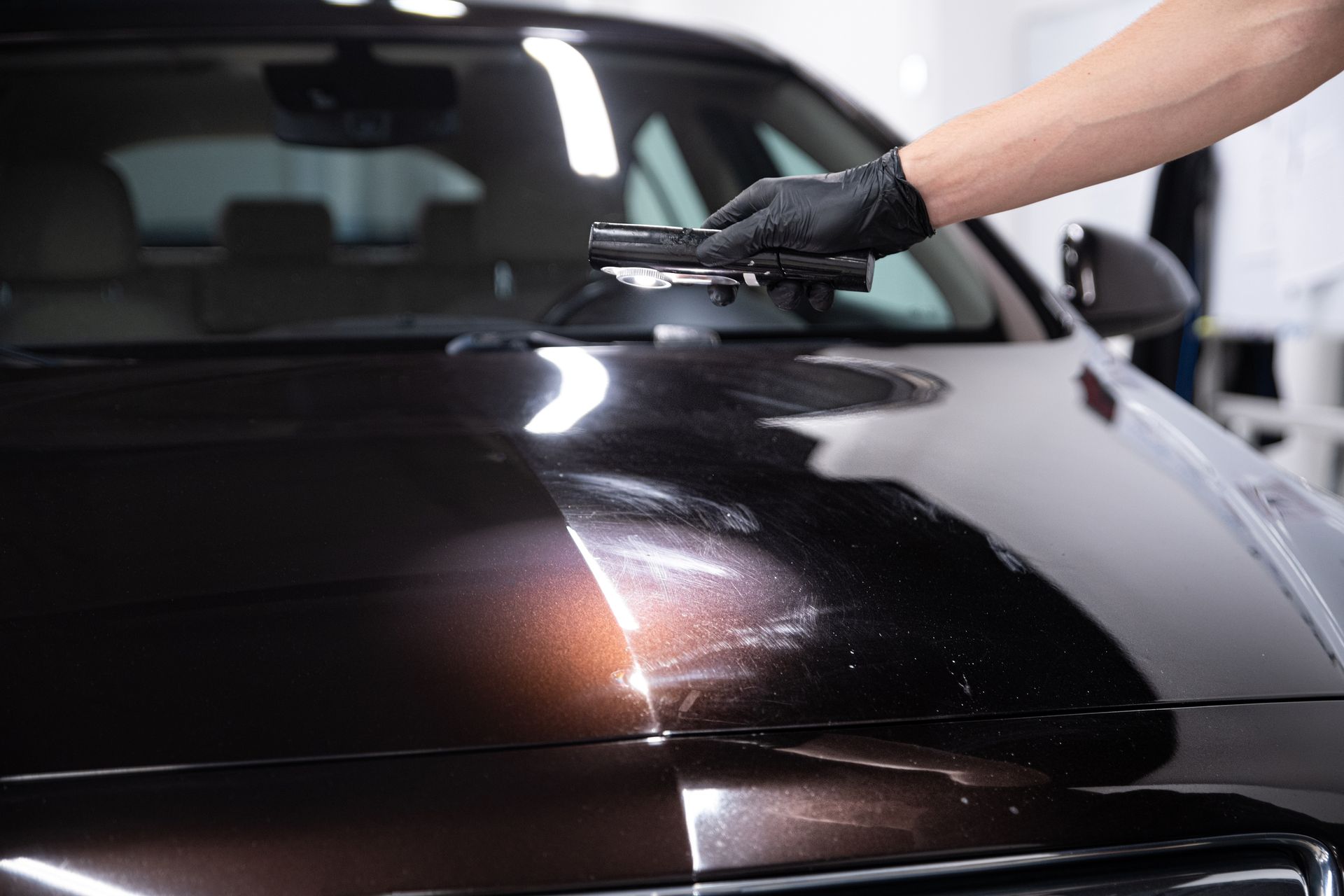 A person in a black glove inspects a car hood with a light, showing a contrast between polished and unpolished areas.
