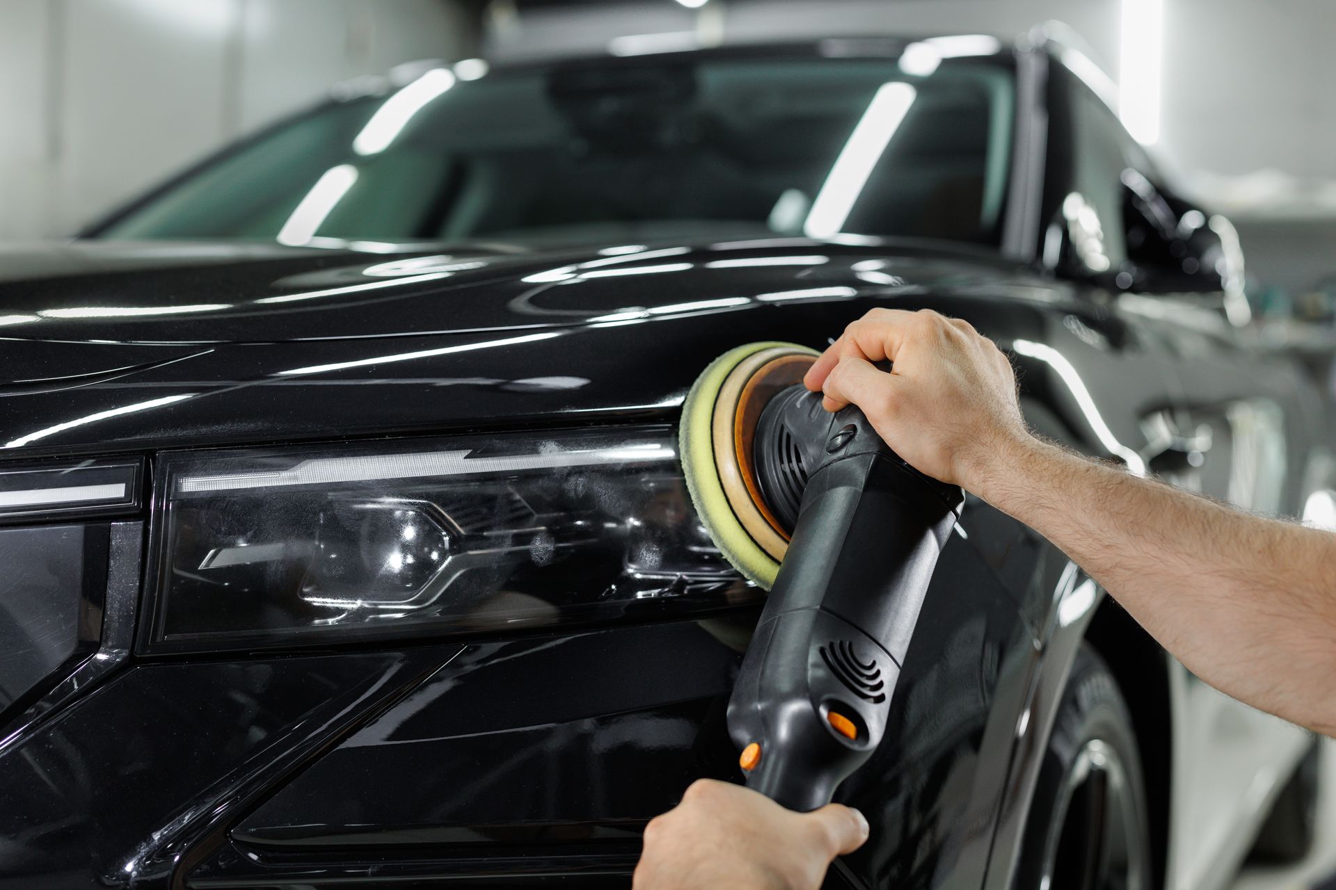 A person uses a power polisher on the headlight and hood of a black car in a garage.