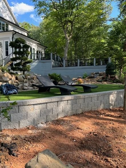 Landscaped backyard with stone wall, sun loungers, patio steps, and trees beside a house