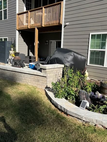 Backyard patio with a grill, covered furniture, stone retaining wall, and upstairs balcony on a gray house