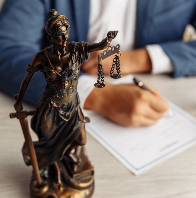 A Statue of Lady Justice on a Table Next to a Man Signing — Budd & Piper Solicitors & Conveyancers in Tweed Heads, NSW