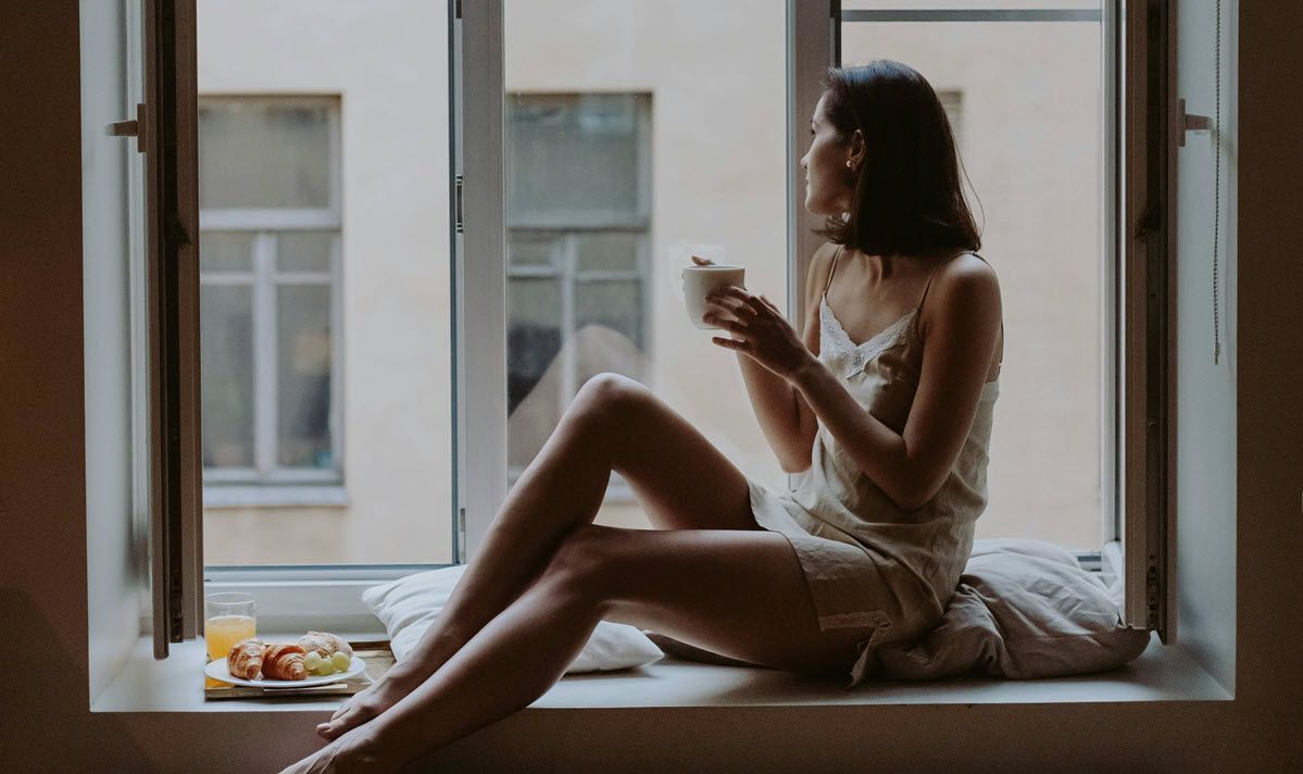 Woman in sleepwear, holding mug, sitting in window, looking outside. A tray with food sits nearby.