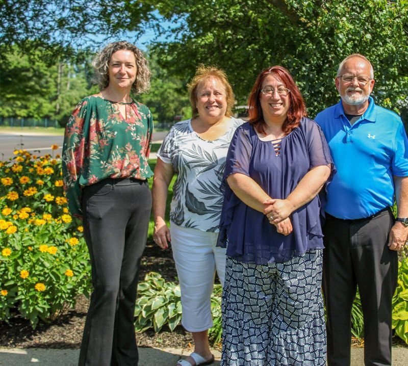 Four people posing outdoors near flowers, trees, and a sidewalk; includes two women, two men.