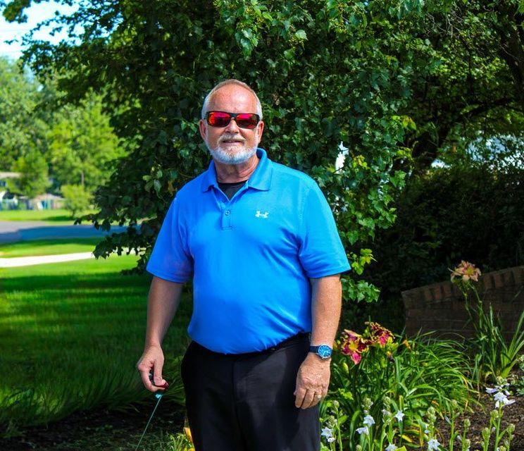 Man in blue shirt and sunglasses smiles outside, next to flowers.