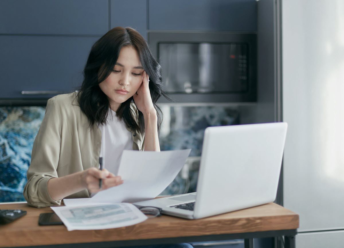 Woman at a desk, looking at documents with a laptop open. She holds a pen, appearing concerned.