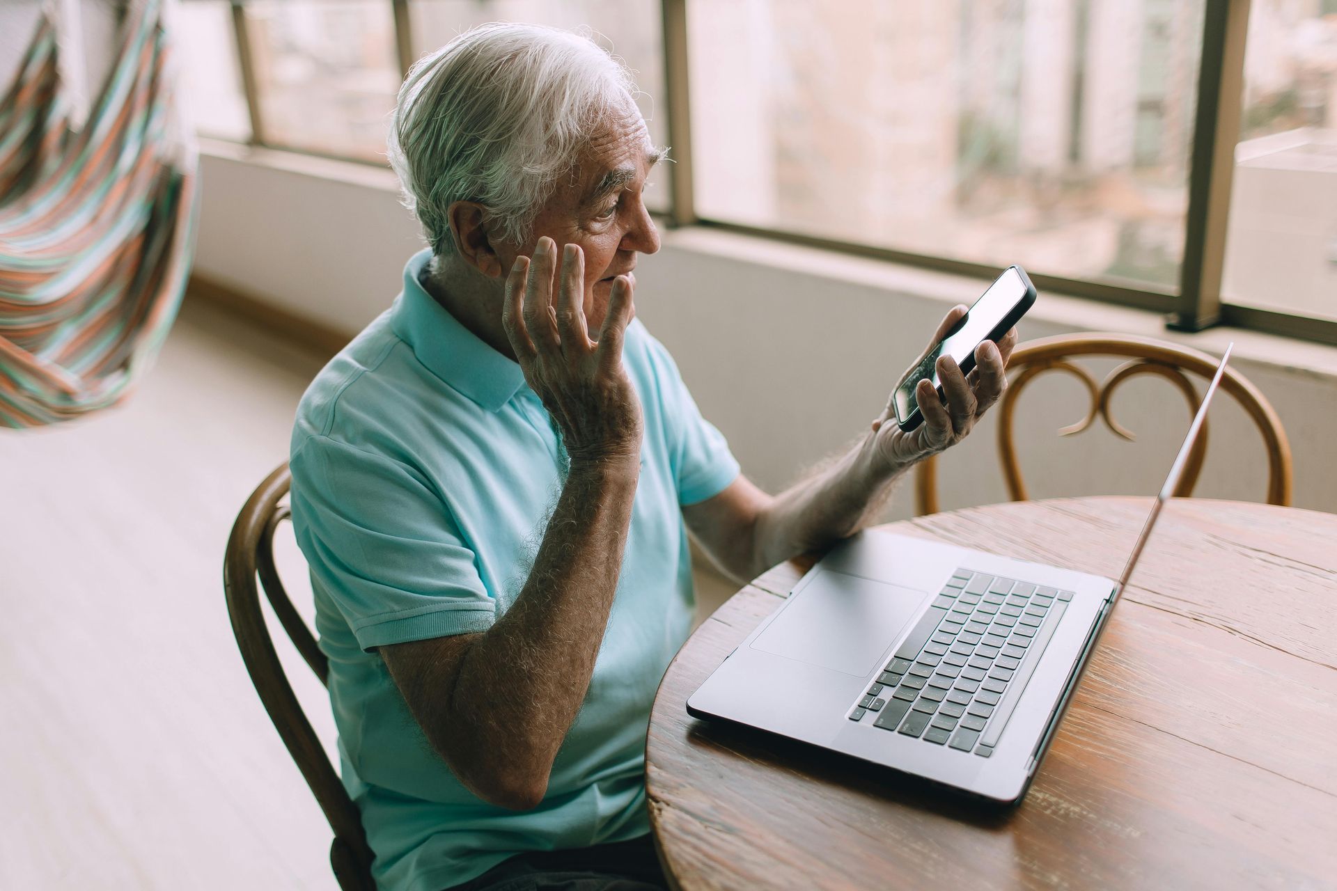 Elderly man in a blue shirt using a smartphone and laptop at a round wooden table, looking at the phone with a smile.