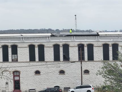 White brick building with boarded-up windows. A person in a yellow vest works on the roof.