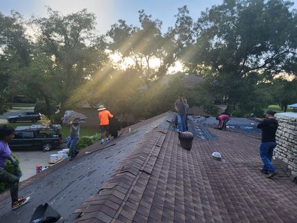 Workers on a roof removing shingles, sun shining through trees in the background.