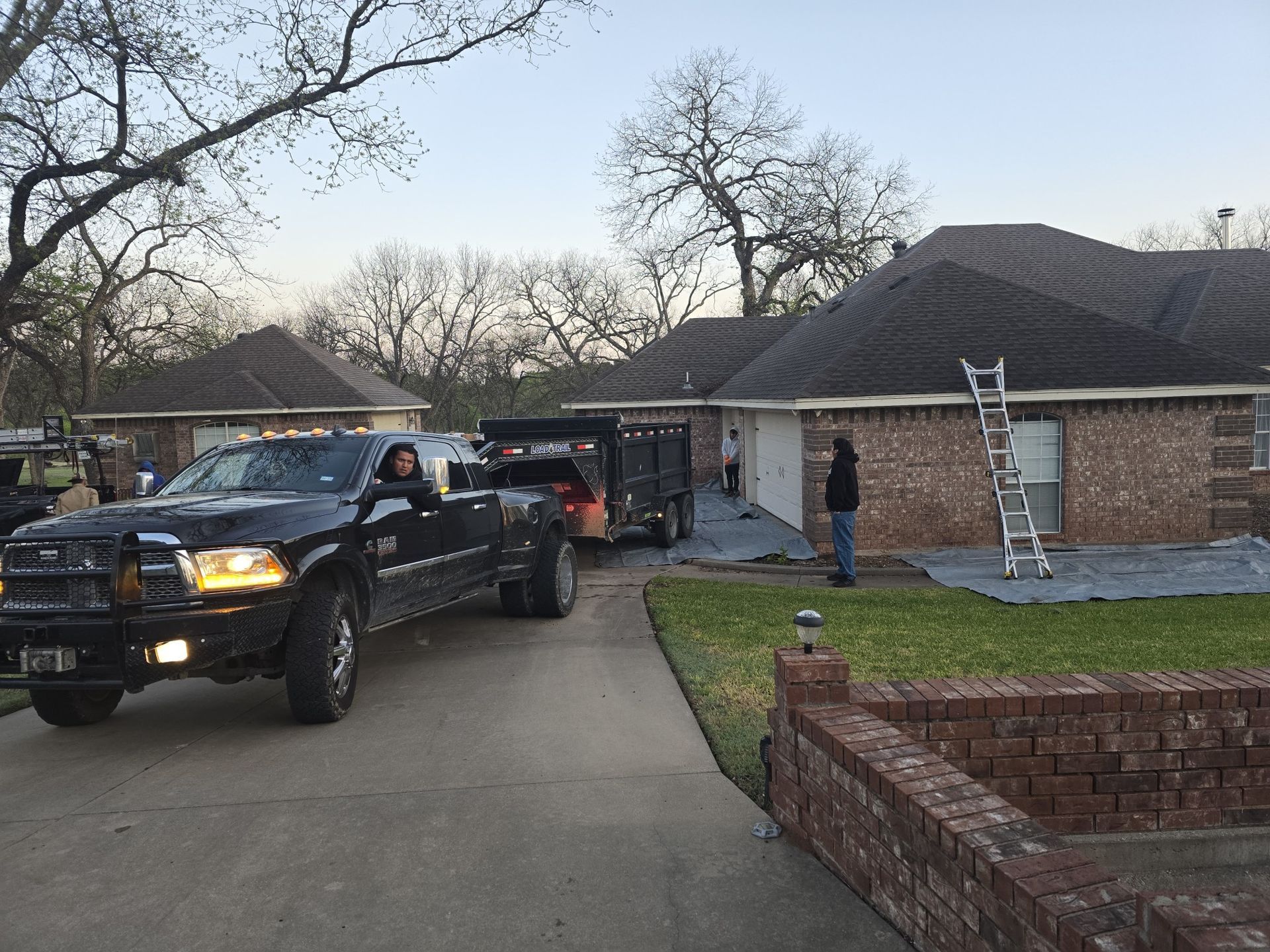 A black truck pulling a trailer in front of a brick house; workers, ladder, and roofing materials are visible.