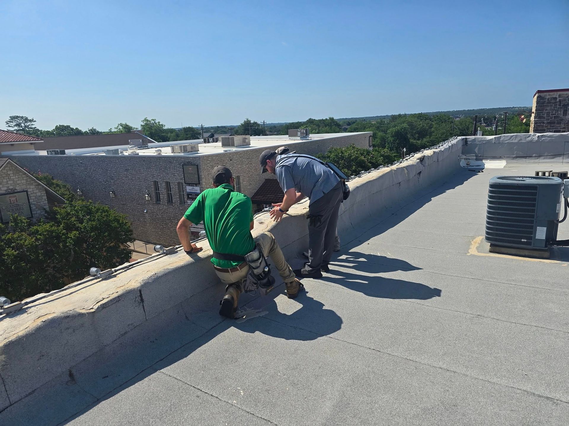 Two men on a flat rooftop, one helping the other climb over a low wall. Sunny day.