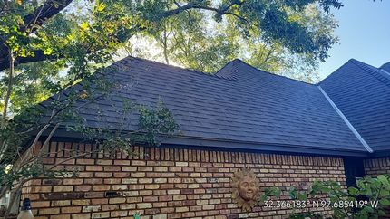 Brick house with dark shingle roof against a backdrop of trees.