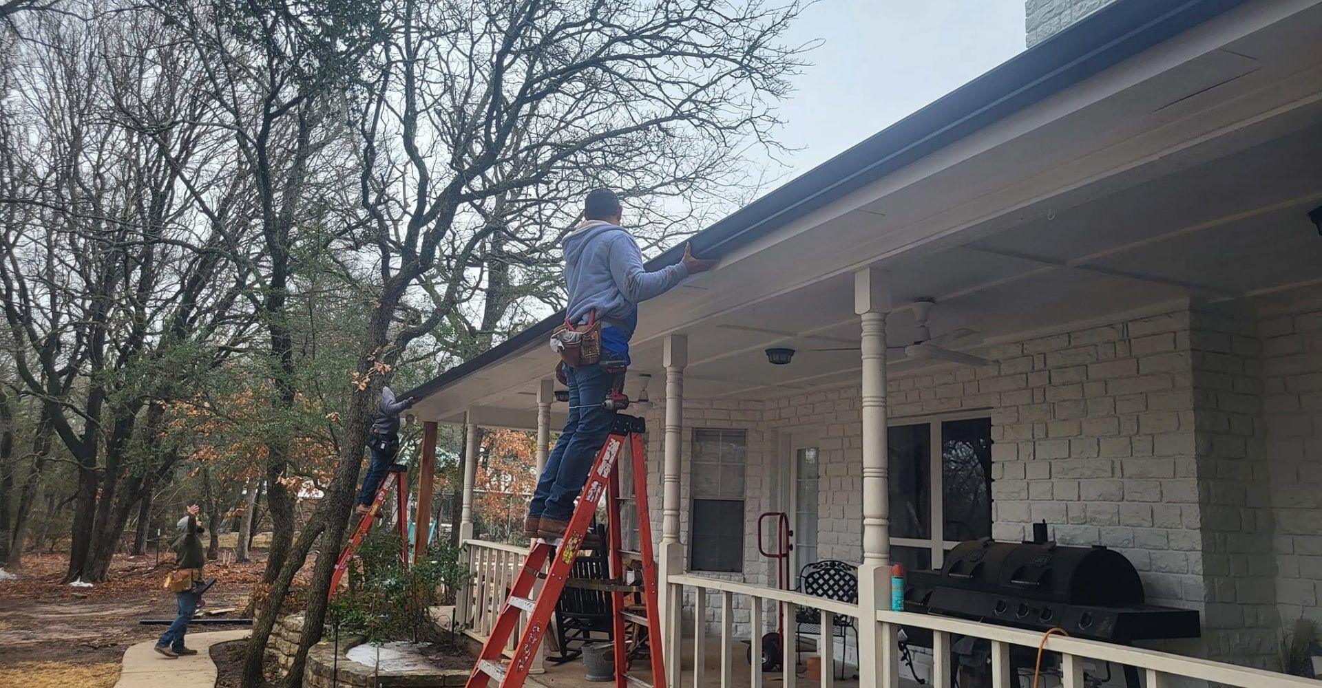 Three people on ladders installing gutters on a white house with a porch, set in a wooded area.