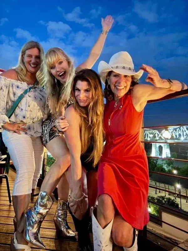 Four women posing and dancing on a rooftop at night, one in a red dress and white cowboy hat