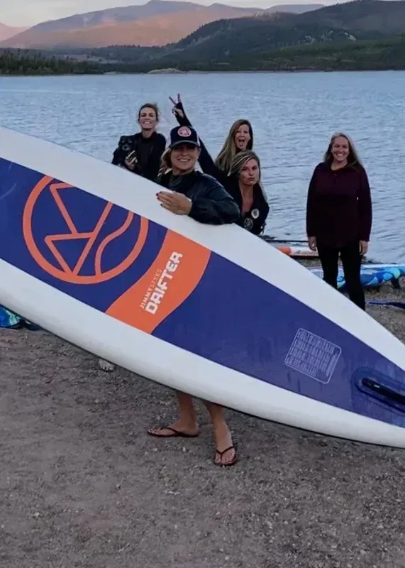 Group of people behind a large paddleboard by a lake at dusk, with mountains in the background.