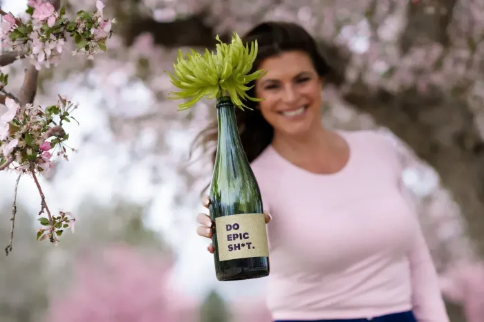 Person holding a green bottle with flowers and a “do epic shit” label, smiling outdoors among blossoms