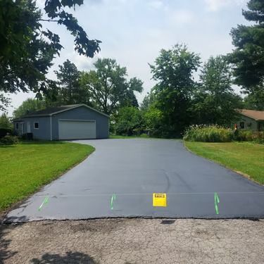 Freshly paved driveway leading to a gray garage and house, with green lawn and trees on both sides.
