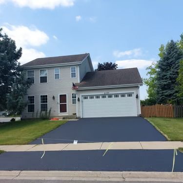Suburban two-story house with attached garage, dark driveway, and a wooden fence under a partly cloudy sky
