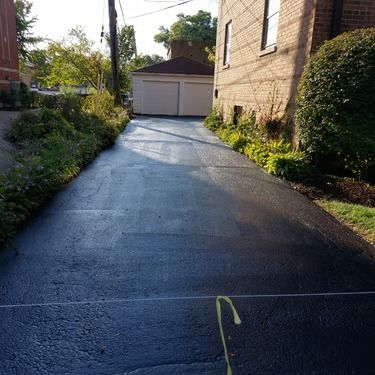 Sloped driveway leading to a garage, bordered by greenery and brick buildings in sunlight.